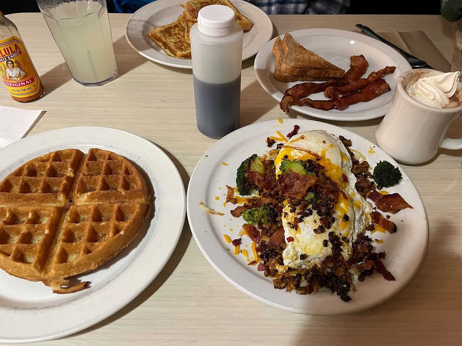 A table with waffles, eggs and vegetables, toast, bacon, a drink, syrup bottle, and a foam coffee.