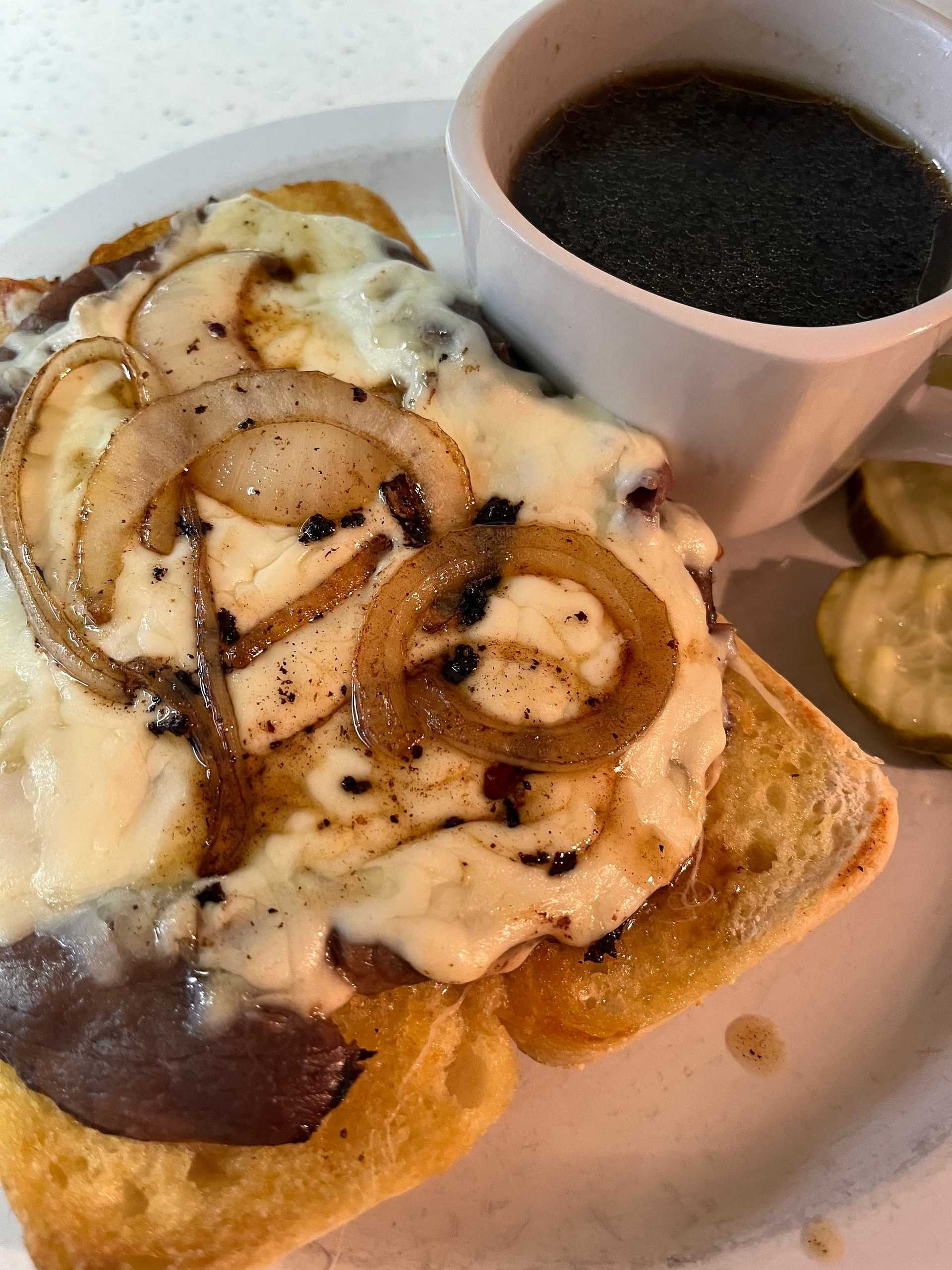 Open-faced sandwich with melted cheese and onions, a cup of black coffee, and sliced pickles.