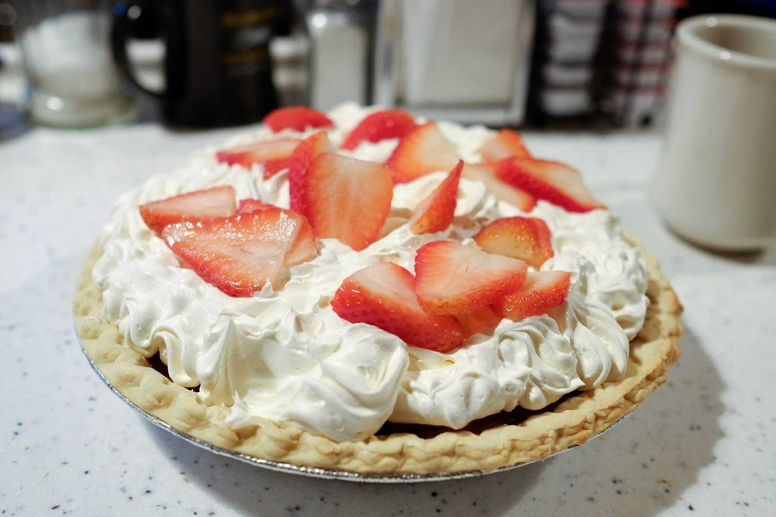 Strawberry-topped cream pie on a kitchen counter, surrounded by a mug and utensils.