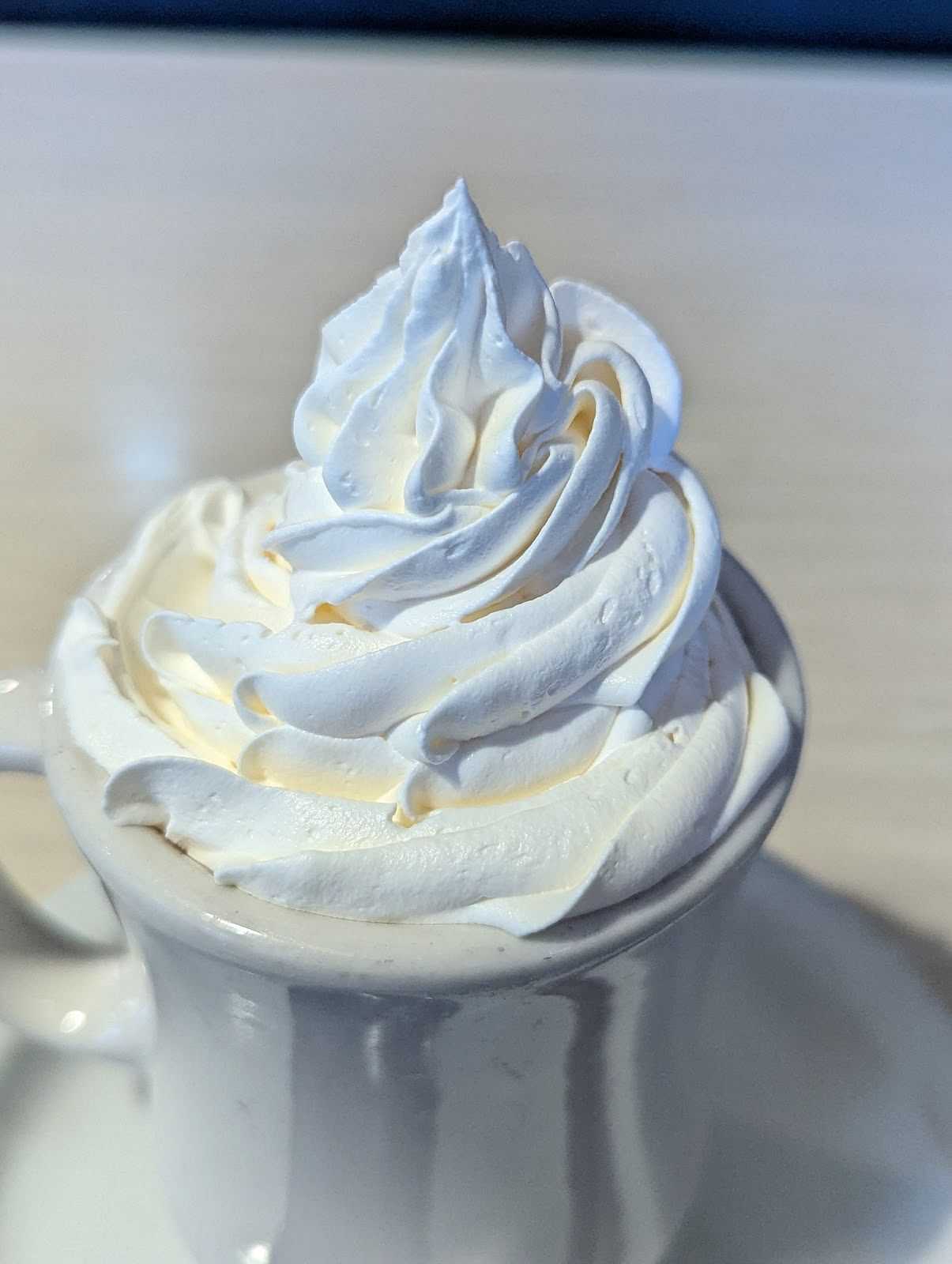 Cup filled with creamy, swirled whipped cream on a saucer, atop a light wooden surface.